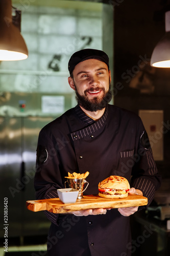 Smiling master chef in uniform looking at camera, holding plate with served burger.