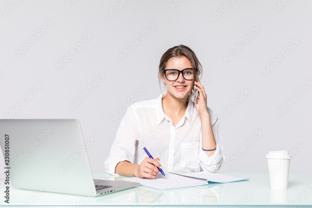 Beautiful businesswoman sitting at the desk with coffee cup, make notice and talking on the phone isolated on the white background Beautiful businesswoman sitting at the desk with coffee cup, make notice and talking on the phone isolated on the white background