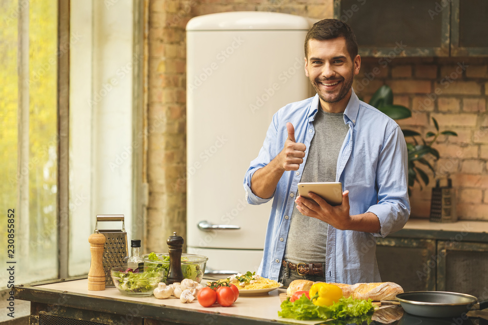 Portrait of young man preparing delicious and healthy food in the home kitchen on a sunny day, using tablet.