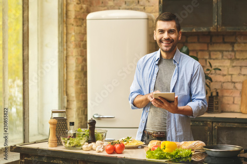 Portrait of young man preparing delicious and healthy food in the home kitchen on a sunny day  using tablet.