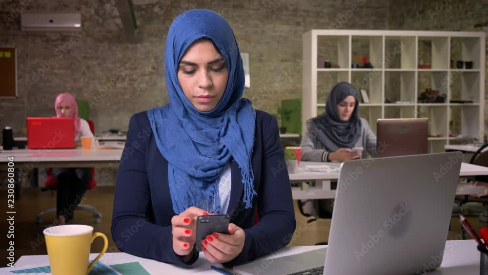 Serious face of amuslim female in blue beutiful hijab is scrolling her phone while sitting in modern studio, surrounded with other working arab girls Serious face of amuslim female in blue beutiful hijab is scrolling her phone while sitting in modern studio, surrounded with other working arab girls