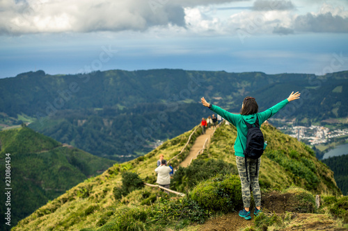 Young woman in khaki clothing is standing on the most popular place on Azores islands - Miradouro da Boca do Inferno. Backside view and raised up hands. Portugal, Sao Miguel.