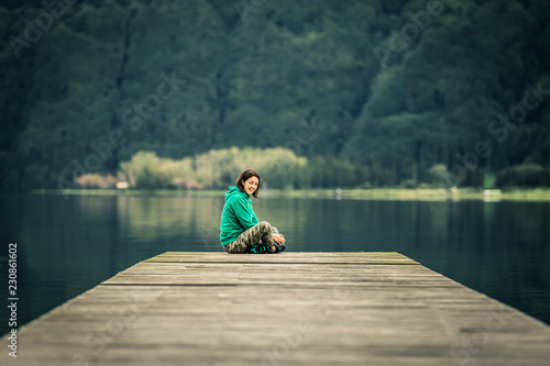 Portrait of young woman in green sweatshirt sitting on the edge of pier. There is volcanic lake on the background. Peaceful atmosphere. Smiling and looking at camera. Azores islands, Portugal