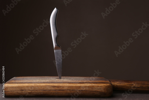 Knife stuck in cutting board on dark background. Wood cutting board and kitchen knife against dark brown wall with copy space.