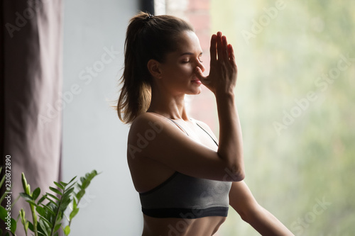 Young attractive woman practicing yoga, doing Alternate Nostril Breathing exercise, nadi shodhana pranayama pose, working out, wearing sportswear, grey top, indoor close up, yoga studio, side view