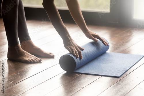 Equipment for sport training session concept. Girl wearing grey sporty pants rolling fitness mat before or after working out in yoga studio club or at home on wooden floor. Hands and legs close up photo