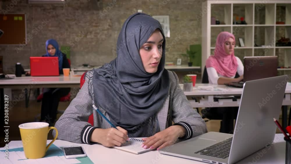 Amazing islamic girl is making notes and looking calmly at her computer, while sitting at desktop, arabic girls on background
