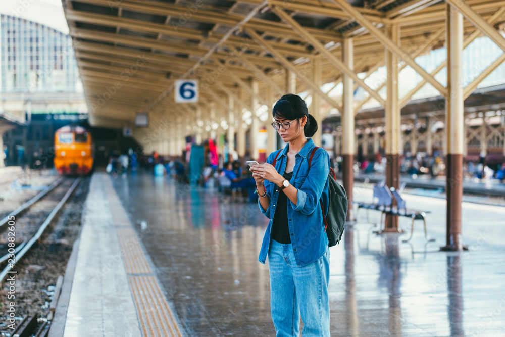 Asian woman traveler used smart phone while wait and check train schedule on the platform of the railway station - travel and transportation concept