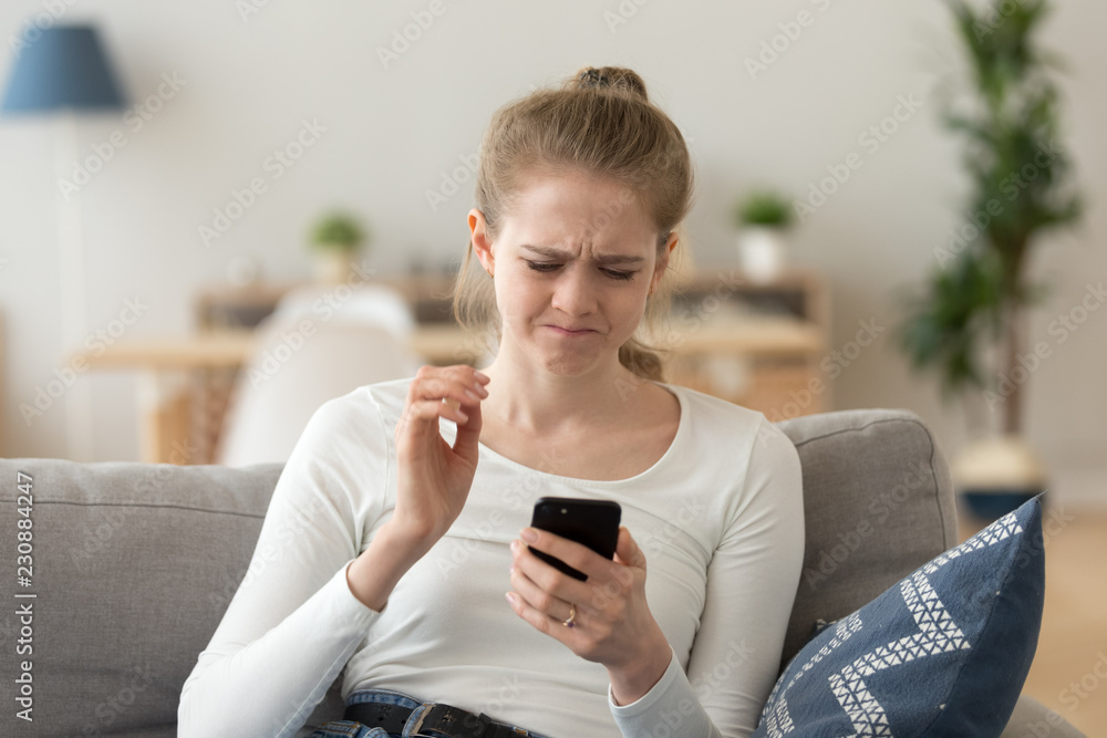 Frustrated young woman sitting alone on couch at home holding looking at discharged or broken mobile phone. Worried female having verification passwords problems wrong combination or received bad news Frustrated young woman sitting alone on couch at home holding looking at discharged or broken mobile phone. Worried female having verification passwords problems wrong combination or received bad news
