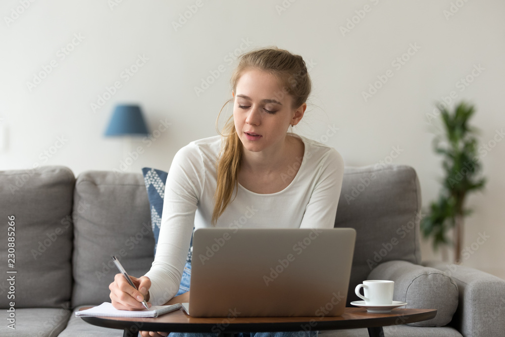 Young millennial woman sitting on couch in room using computer making notes on exercise book working or studying alone at home. Concentrated student preparing for test or examination learning indoors Young millennial woman sitting on couch in room using computer making notes on exercise book working or studying alone at home. Concentrated student preparing for test or examination learning indoors