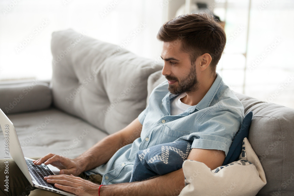 Young millennial positive man sitting on couch in living room with laptop alone on weekend at home. Handsome male using notebook have fun looking recreational video or freelancer working online indoor