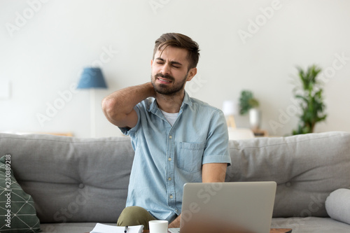 Close up millennial man sitting on couch touching massaging neck. Exhausted tired male suffering from neck pain ache after sedentary work or study at home for a long time feeling unwell and discomfort photo