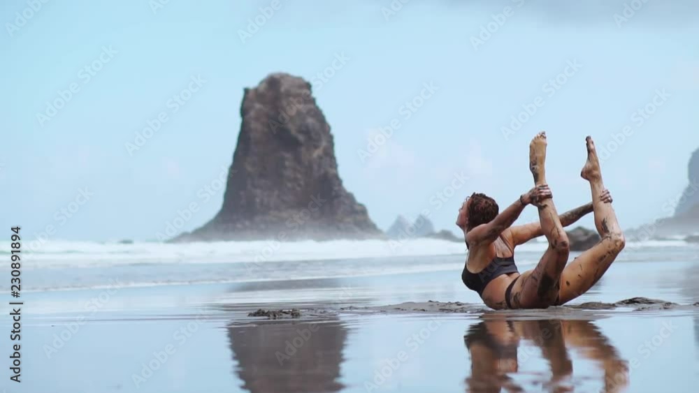 A beautiful woman in a swimsuit performs an exercise bridge on the beach with black volcanic sand surrounded by mountains and ocean. Gymnastics and yoga A beautiful woman in a swimsuit performs an exercise bridge on the beach with black volcanic sand surrounded by mountains and ocean. Gymnastics and yoga