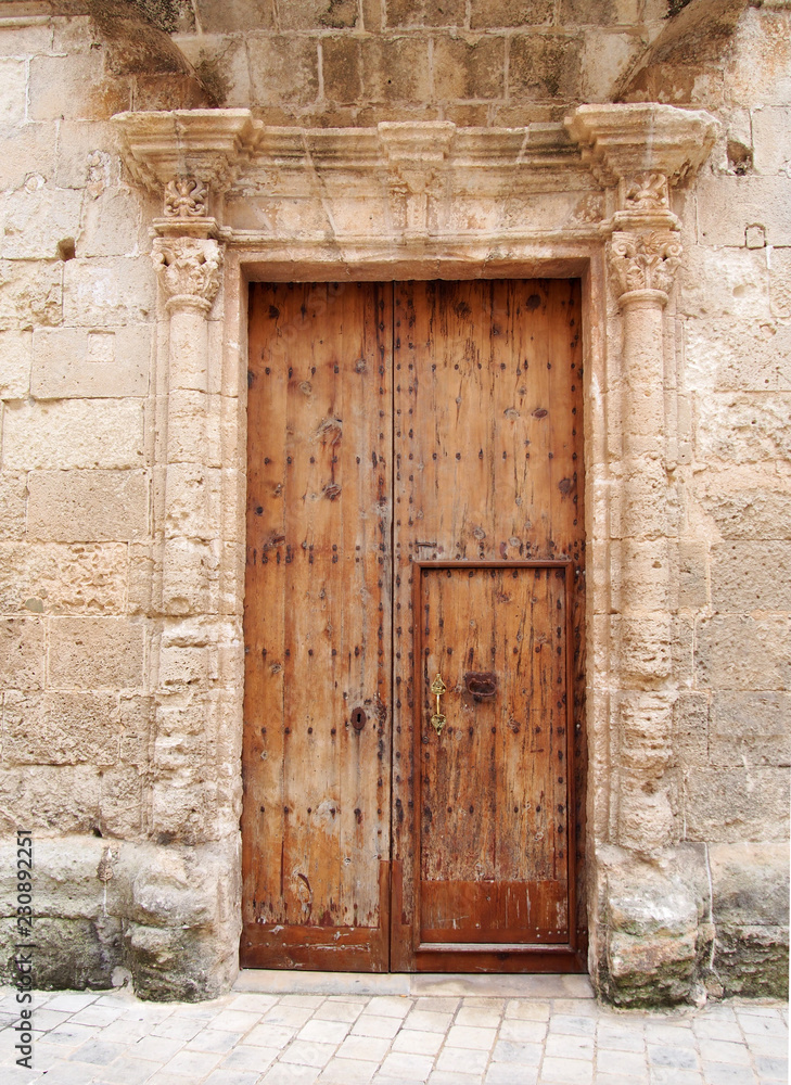 large old brown wooden door covered with rusted iron studs keyhole and handle set in an ornate carved stone frame with surrounding wall