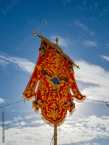 Red beautiful flag for annual festa near church festively decorated square in Malta on blue sky background