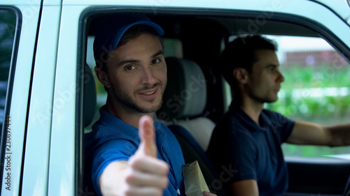 Young delivery man holding parcel, showing thumbs up in van, express shipment
