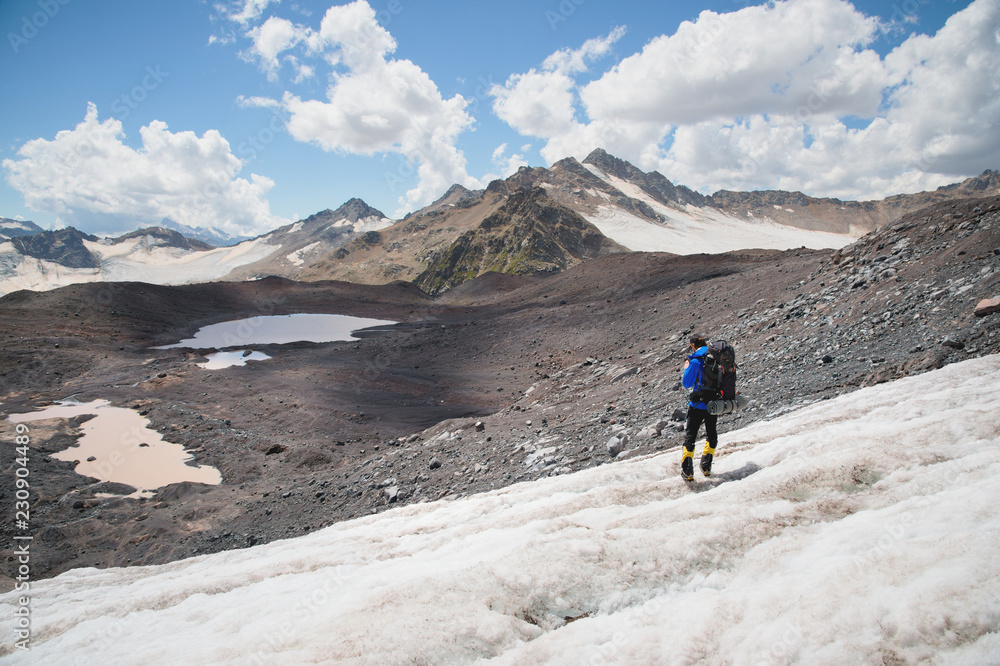 Traveler in a cap and sunglasses with a backpack on his shoulders in the snowy mountains on the glacier against the sky and clouds. Traveler in a natural environment