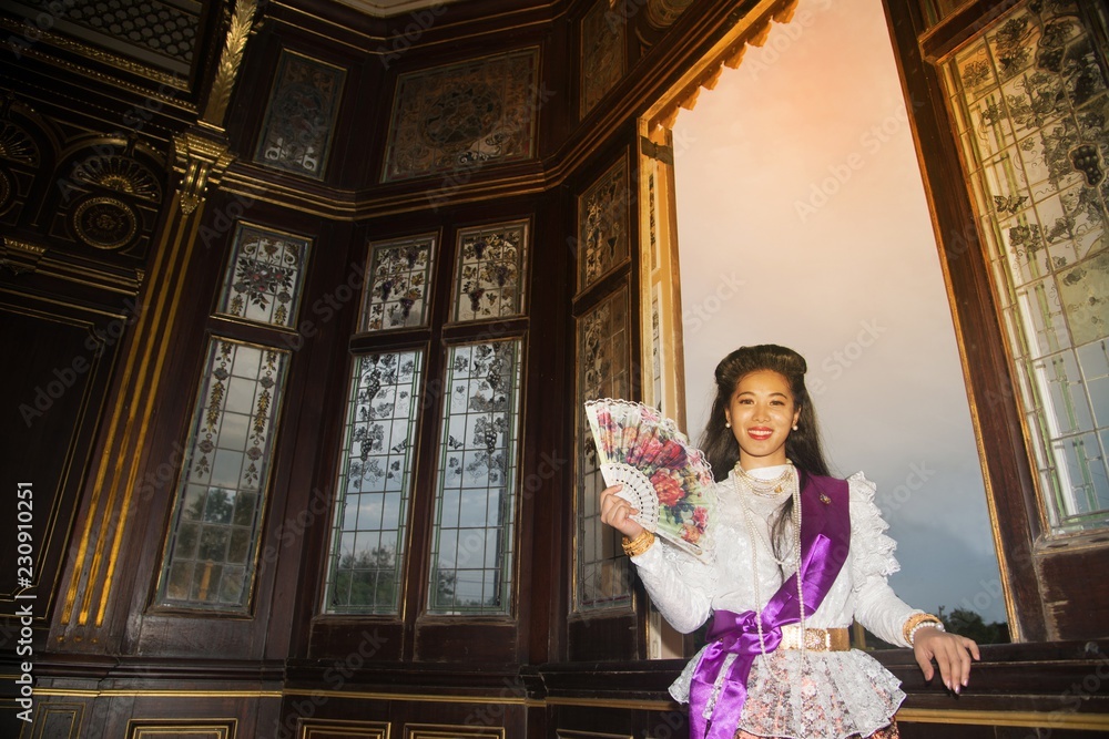 The lady in Middle Thai classical Thai traditional dress suit and hold a paper fan is posing standing indoor at window of ancient 
railway station building.