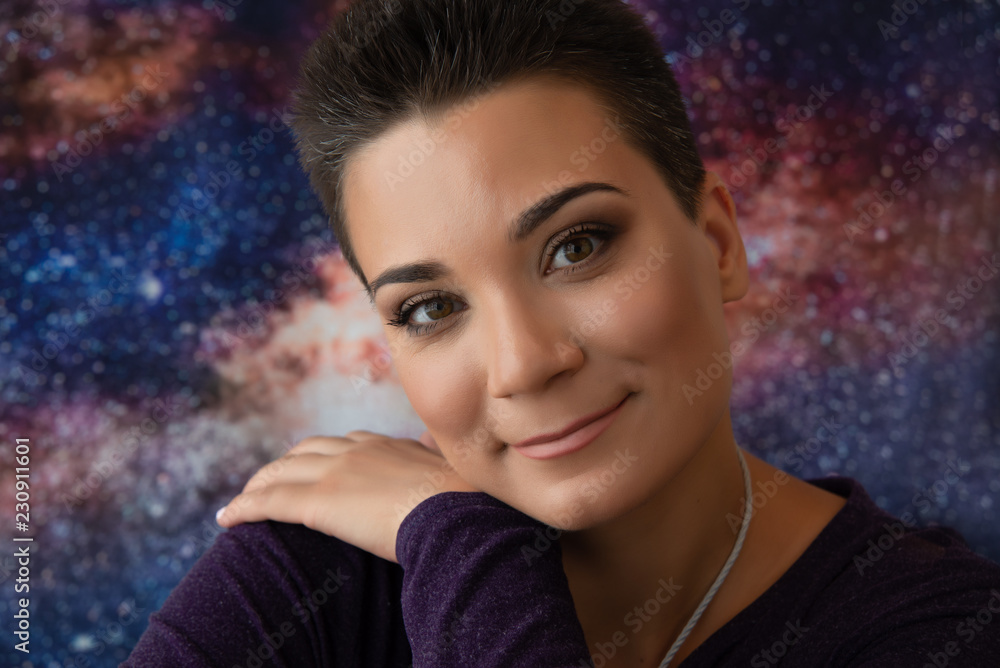 A young woman astrologer with smooth skin and short, striped hair smiles and puts her hand on her shoulder. Photo on the cosmic space background. A young woman astrologer with smooth skin and short, striped hair smiles and puts her hand on her shoulder. Photo on the cosmic space background.