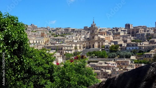 Modica, Sicily - Italy landscape with the catholic church (Cathedral of San Giorgio) front view. Old modica houses around the church. Leaves of the top of a tree sway in the foreground.