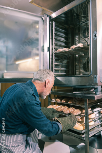 Early morning. Bearded owner of bakery putting croissants into oven working early in the morning