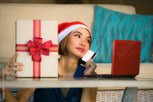 young happy and beautiful girl relaxed at home couch in Santa hat using laptop computer paying for Christmas present with credit card smiling in online shopping