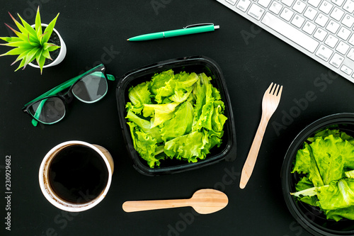 Order healthy food delivery to office. Light, diet meal with lettuce in plastic containers, coffee, disposable spoon and fork near computer keayboard and glasses on black background top view photo