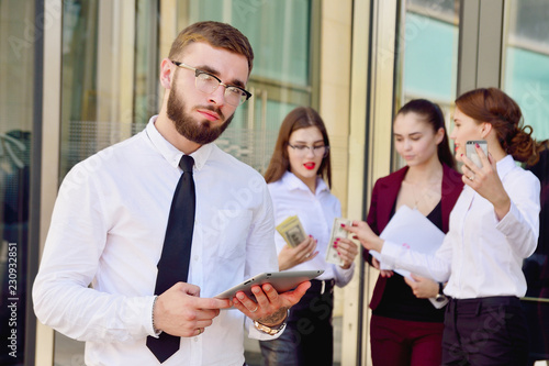 A young male manager with a tablet in his hands against the background of the girls. Office staff in the background of a multi-storey glass building. Lifestyle. Businessman