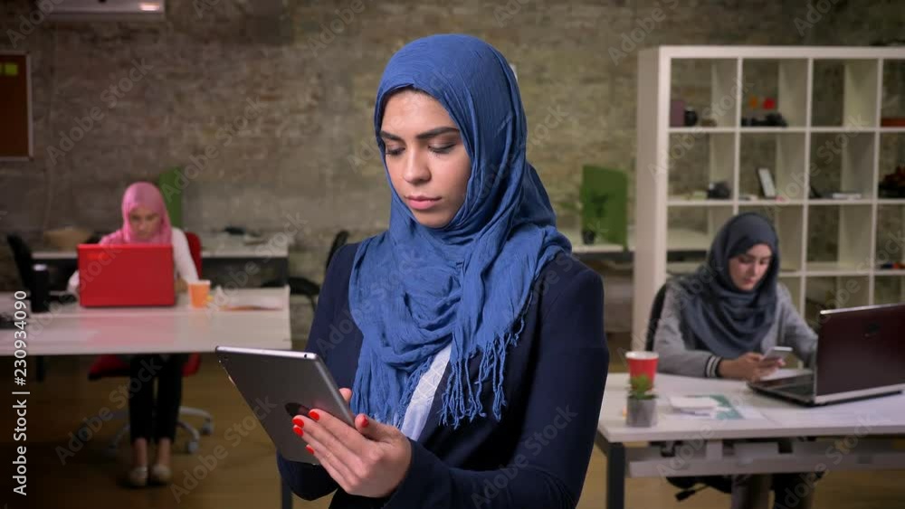Serious focused arab female in blue hijab standing and typing on her tablet, arabic females on background, modern illustration indoor