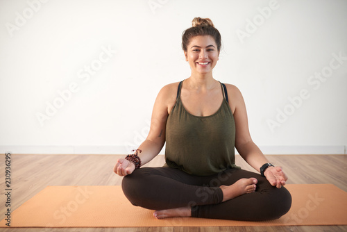 Joyful young plus-size woman sitting in lotus position on yoga mat