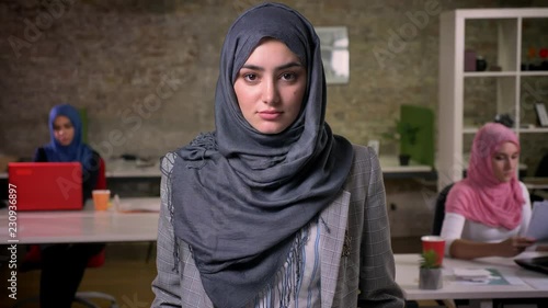 Beautiful arab woman in grey hijab is standing quietly and calmly looking at camera, brick background, working colleagues ar sitting and using theis laptops photo