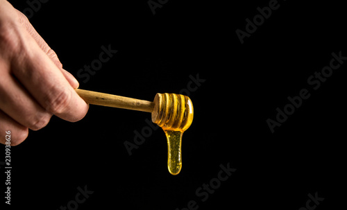 Man hand holding wooden tuned wood spoon dipper on top of small transparent glass jar, dripping liquid honey, tasty golden yellow. Brown wooden table against black background.