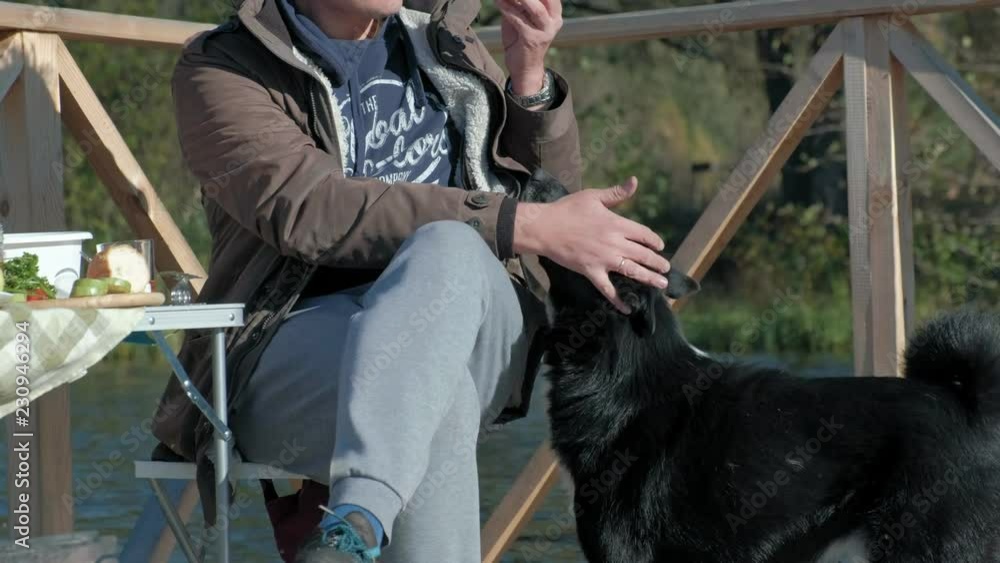mature man in warm clothes, sitting at the table, eating a burger, near a hungry dog, having a picnic on the river bank on a wooden bridge, weekend, cold weather, camping, tourism mature man in warm clothes, sitting at the table, eating a burger, near a hungry dog, having a picnic on the river bank on a wooden bridge, weekend, cold weather, camping, tourism