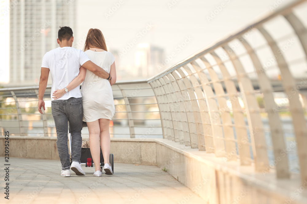 Young couple walking along bridge with stroller, view from the back