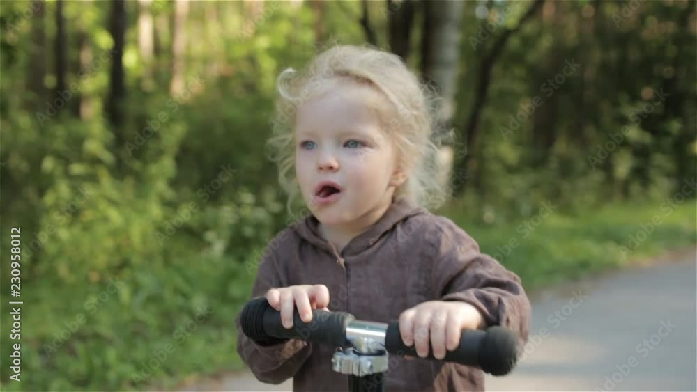 little girl blonde in a brown linen jumpsuit rolls on a scooter on an asphalt path in a pine forest little girl blonde in a brown linen jumpsuit rolls on a scooter on an asphalt path in a pine forest