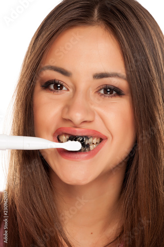 young woman brushing her teeth with electric tooth brush with black active charcoal toothpaste on white background
