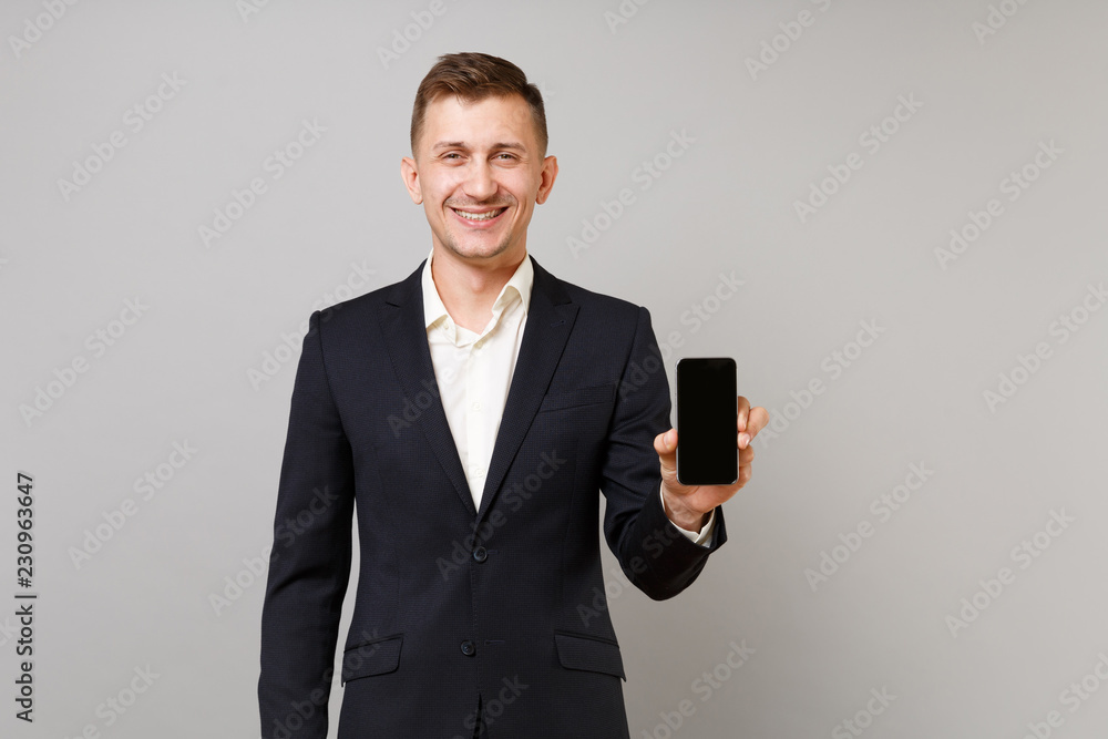 Joyful young business man in classic black suit and shirt holding mobile phone with blank empty screen isolated on grey wall background. Achievement career wealth business concept. Mock up copy space. Joyful young business man in classic black suit and shirt holding mobile phone with blank empty screen isolated on grey wall background. Achievement career wealth business concept. Mock up copy space.