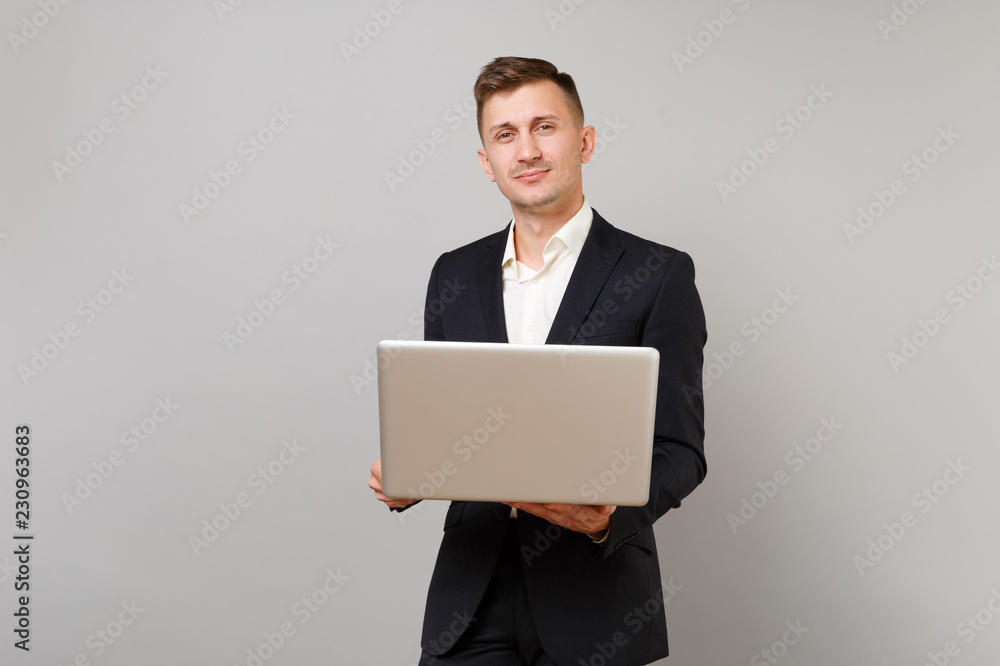 Successful young business man in classic black suit, white shirt holding laptop pc computer isolated on grey wall background in studio. Achievement career wealth business concept. Mock up copy space. Successful young business man in classic black suit, white shirt holding laptop pc computer isolated on grey wall background in studio. Achievement career wealth business concept. Mock up copy space.