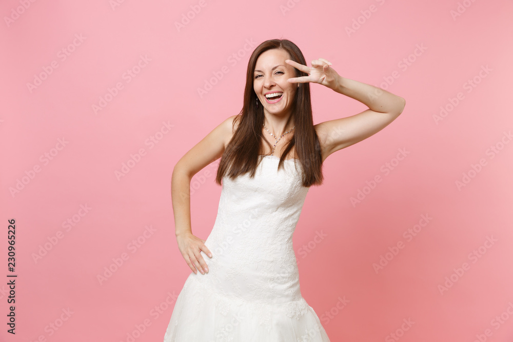 Portrait of laughing bride woman in beautiful white lace wedding dress standing and showing victory sign isolated on pink pastel background. Wedding celebration concept. Copy space for advertisement.