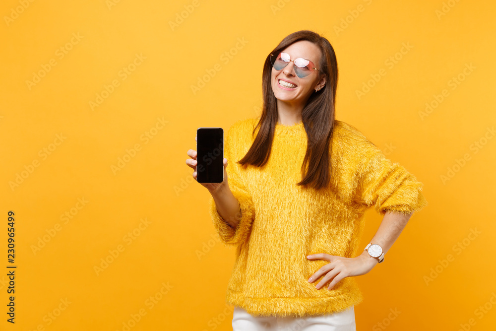 Portrait of smiling young woman in heart glasses holding mobile phone with blank black empty screen isolated on bright yellow background. People sincere emotions, lifestyle concept. Advertising area. Portrait of smiling young woman in heart glasses holding mobile phone with blank black empty screen isolated on bright yellow background. People sincere emotions, lifestyle concept. Advertising area.