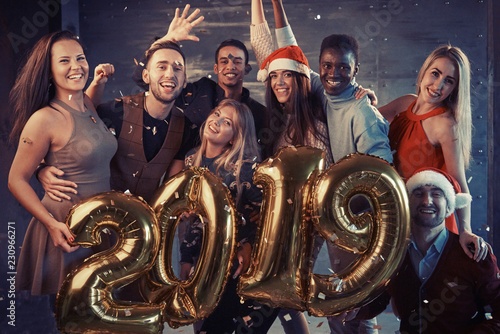 New 2019 Year is coming! Group of cheerful young multiethnic people in Santa hats carrying gold colored numbers and throwing confetti on the party