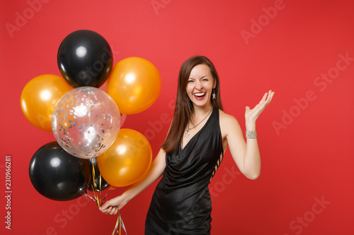 Laughing young girl in little black dress celebrating spreading hands holding air balloons isolated on red background. International Women's Day, Happy New Year, birthday mockup holiday party concept. © ViDi Studio