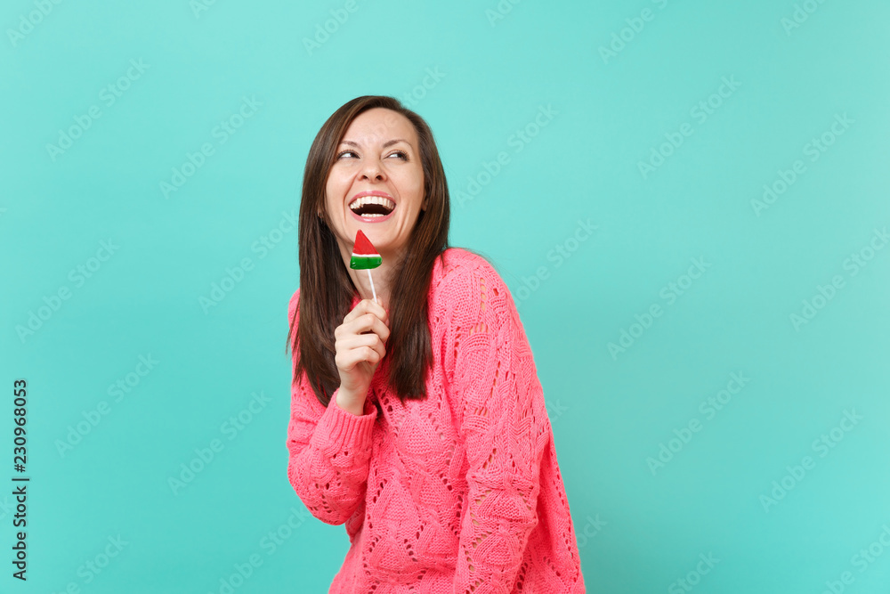 Fototapeta premium Laughing cheery young woman in knitted pink sweater looking up, holding watermelon lollipop isolated on blue turquoise wall background, studio portrait. People lifestyle concept. Mock up copy space.