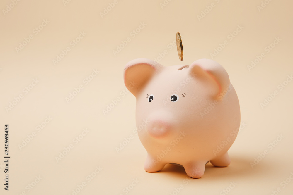 Golden coin falling into pink piggy money bank isolated on pastel beige background. Money accumulation, investment, banking or business services, wealth concept. Copy space advertising mock up. Golden coin falling into pink piggy money bank isolated on pastel beige background. Money accumulation, investment, banking or business services, wealth concept. Copy space advertising mock up.