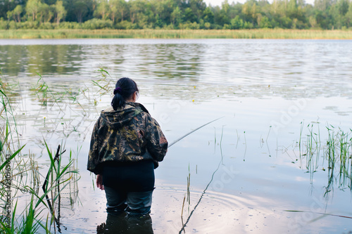 Woman with dark hair in dark clothes and high rubber boots is fishing with a fishing rod on a beautiful lake