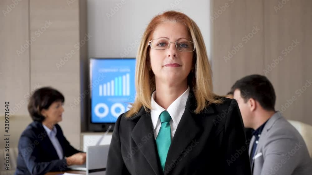 Close up slow motion portrait of smiling businesswoman company CEO in front of the camera while her colleagues are working in the background Close up slow motion portrait of smiling businesswoman company CEO in front of the camera while her colleagues are working in the background