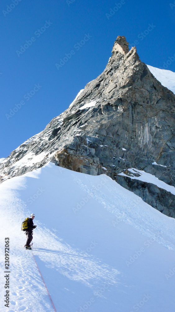 mountain climber standing on a snow and ice ridge below a sharp rock needle mountain peak under a blue sky
