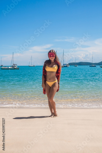 Young smiling woman with long hair in yellow bikini wear funny merry christmas glasses and red tinsel standing on tropical beach with yachts on background, blue water and sky on Phuket island,Thailand