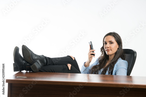 Woman looking at  camera with smartphone in hand  in the office, sitting with legs on desk