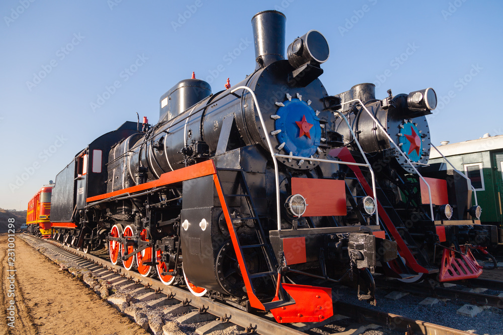 Fototapeta premium Old black, white and red locomotive is standing on the rails in a railway museum against a blue sky. Concept of history, the carriage of passengers, transport
